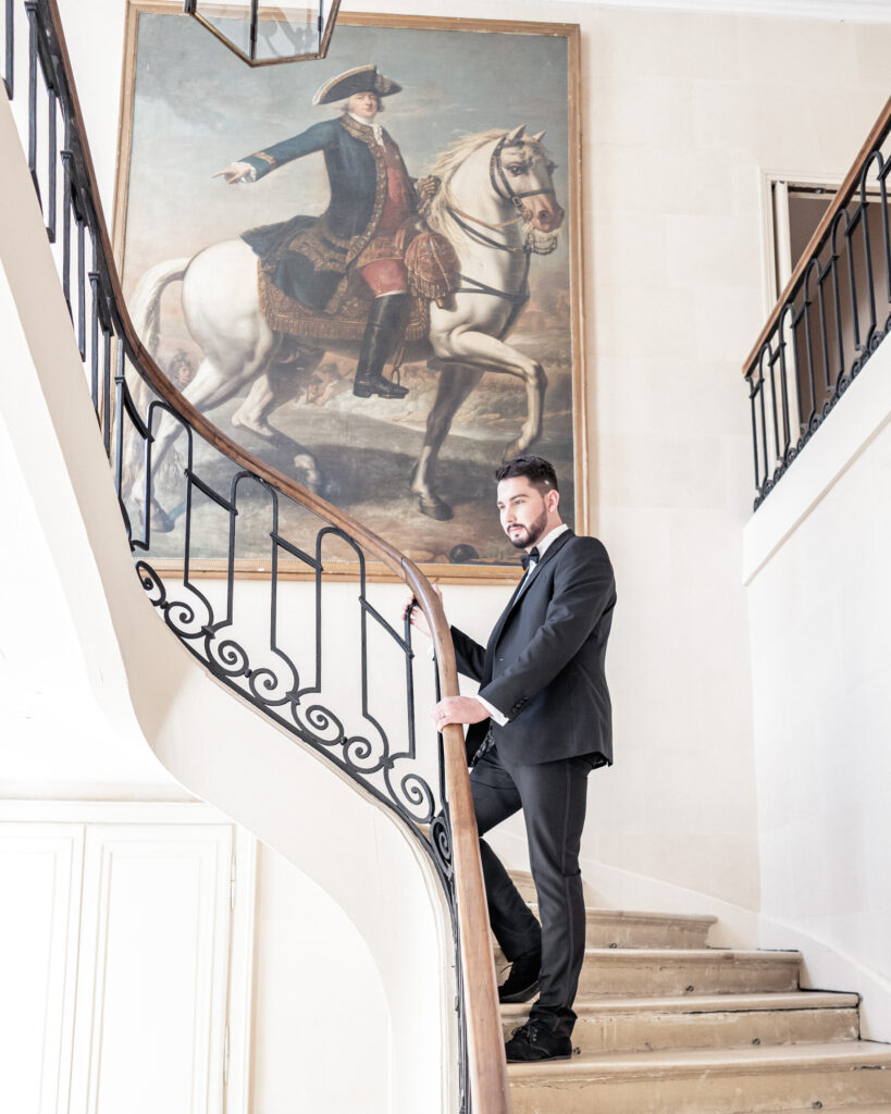 A groom dressed in black tie luxury wedding attire and posing on the grand staircase at Chateau de Courtomer in Normandy, France.