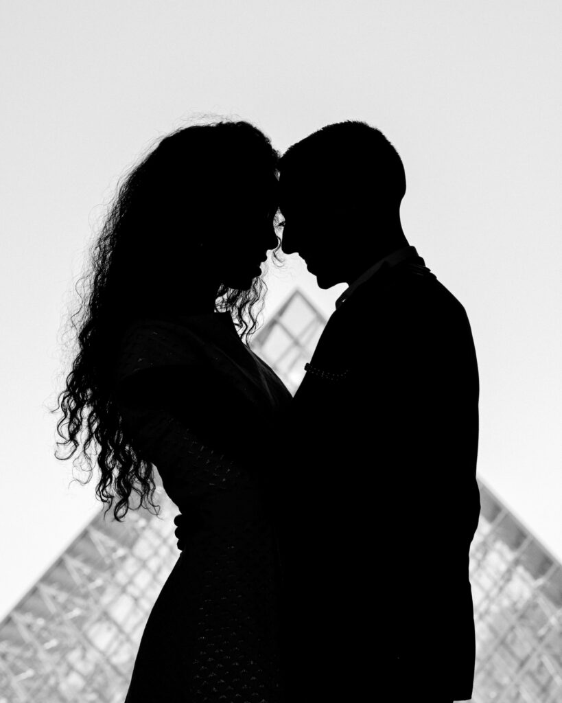 Silhouette of a couple touching foreheads in front of the Louvre museum pyramid.