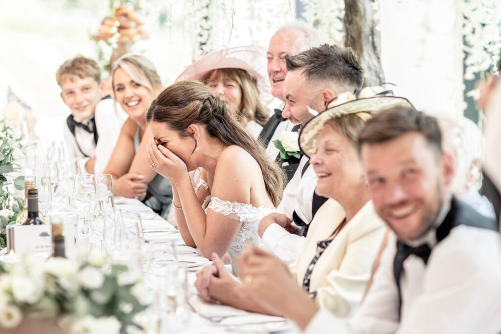 Bride with her head in her hands and laughing as the best man delivers his speech.