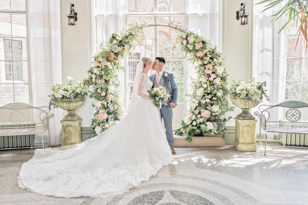 Bride and groom posing together and kissing in front of a fresh floral archway in the winter garden at Hawkstone Hall in England.