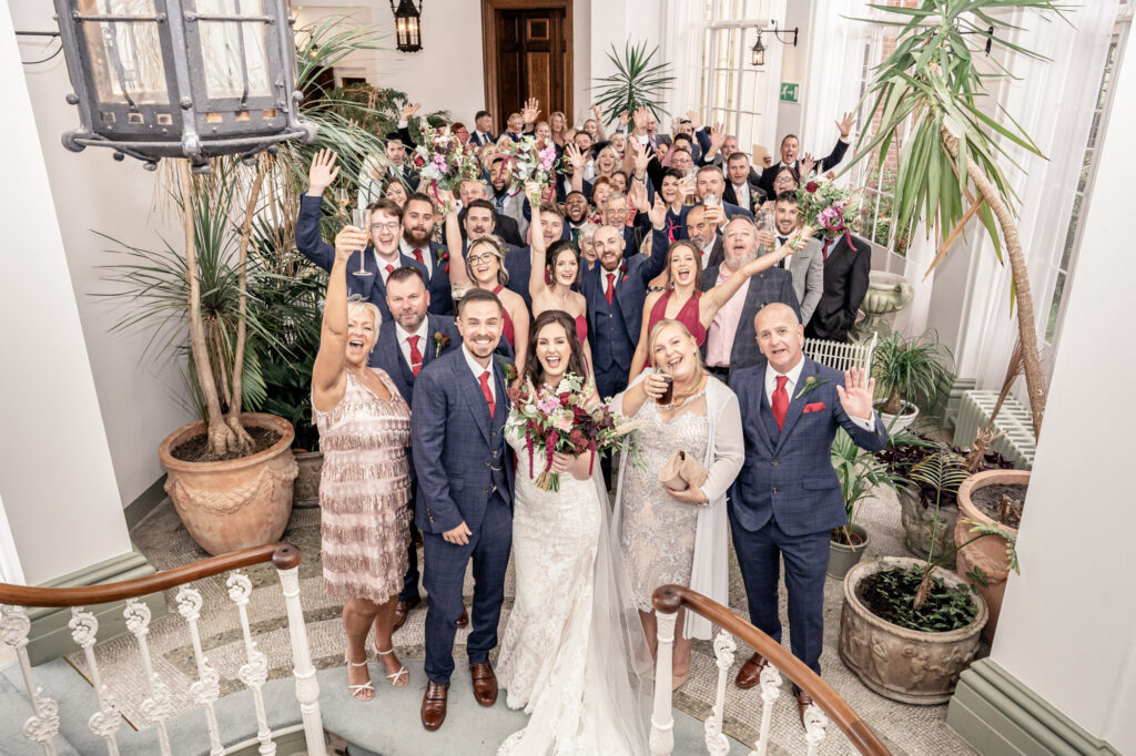 Large group photo of the bride and groom and all wedding guests in the winter garden room at Hawkstone Hall. All guests have their hands in the air cheering and smiling.