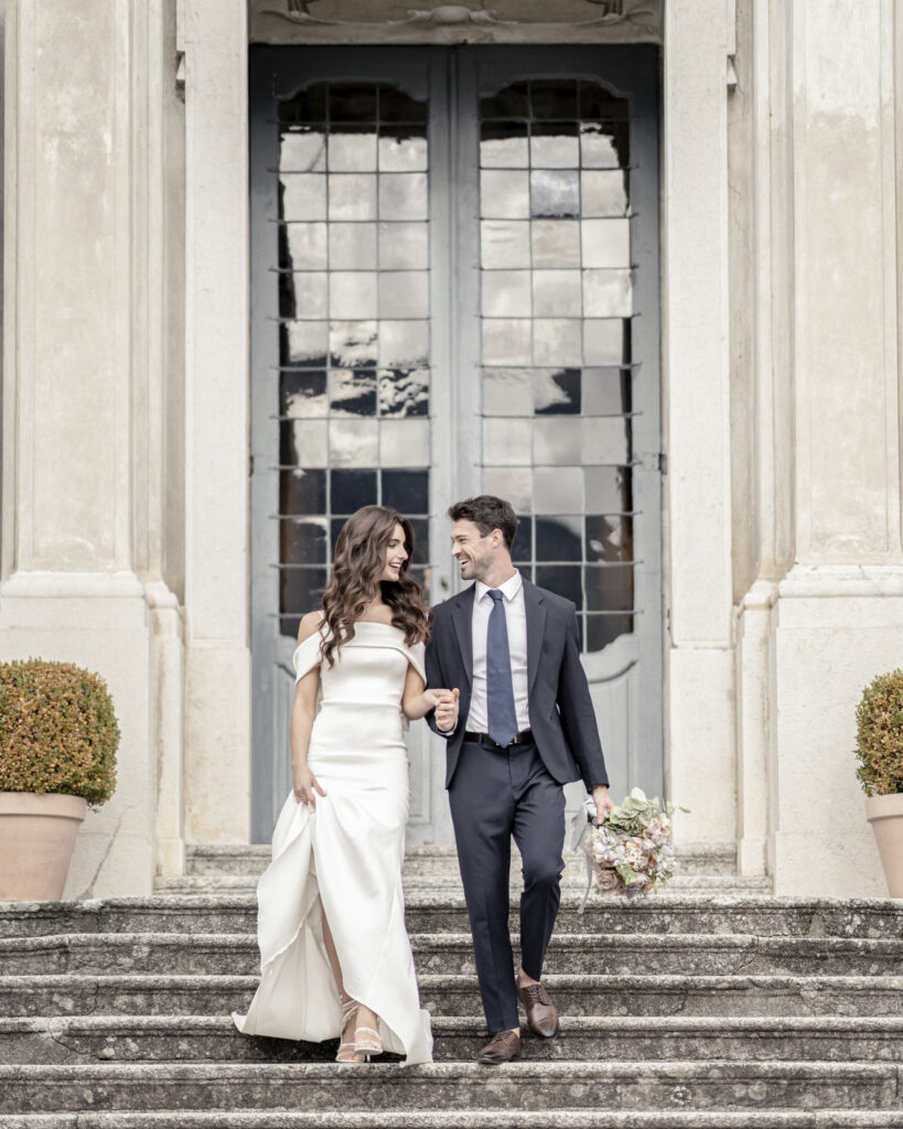 Bride and groom walking down the front steps at Villa Sola Cabiati next to Lake Como, hand-in-hand and smiling.