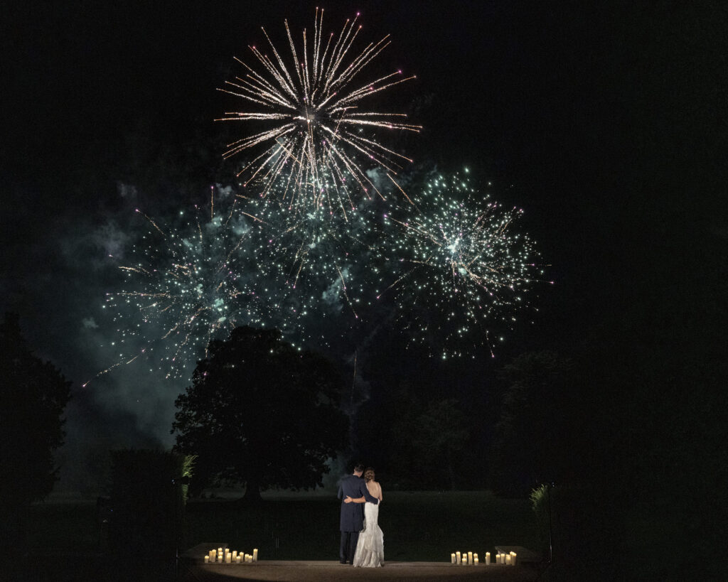 Bride and groom with their arms around each other watching fireworks on their wedding day.