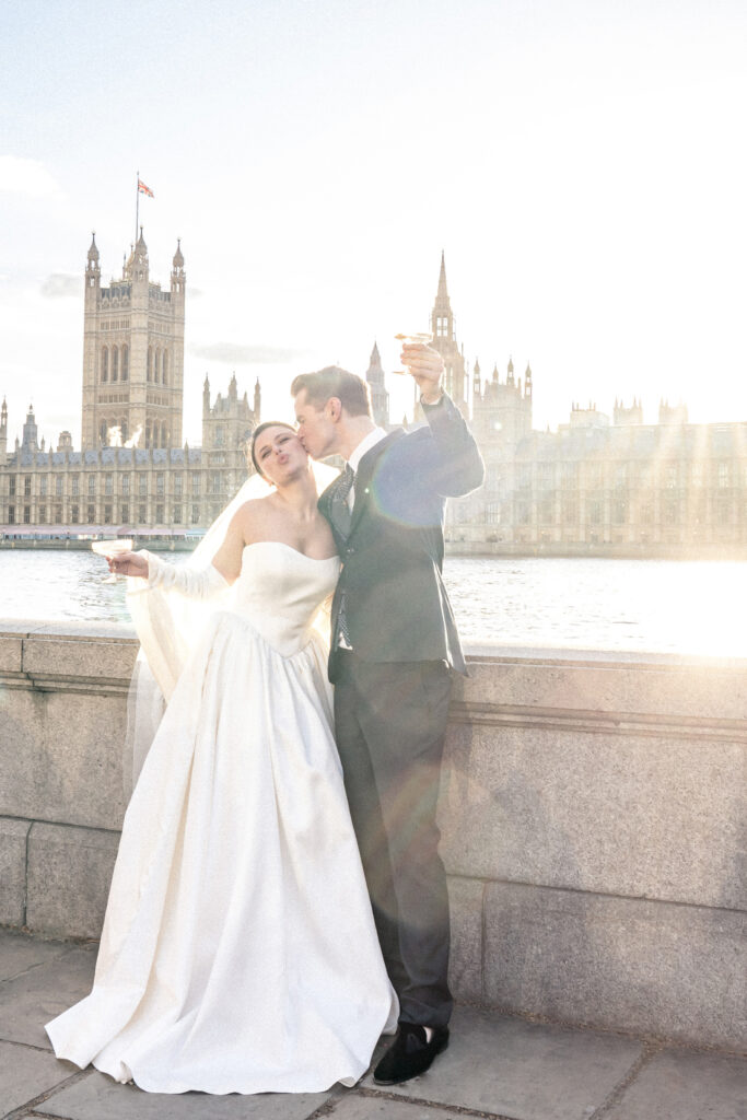 Bride and groom with champagne glasses raising a celebratory toast by the River Thames in London.