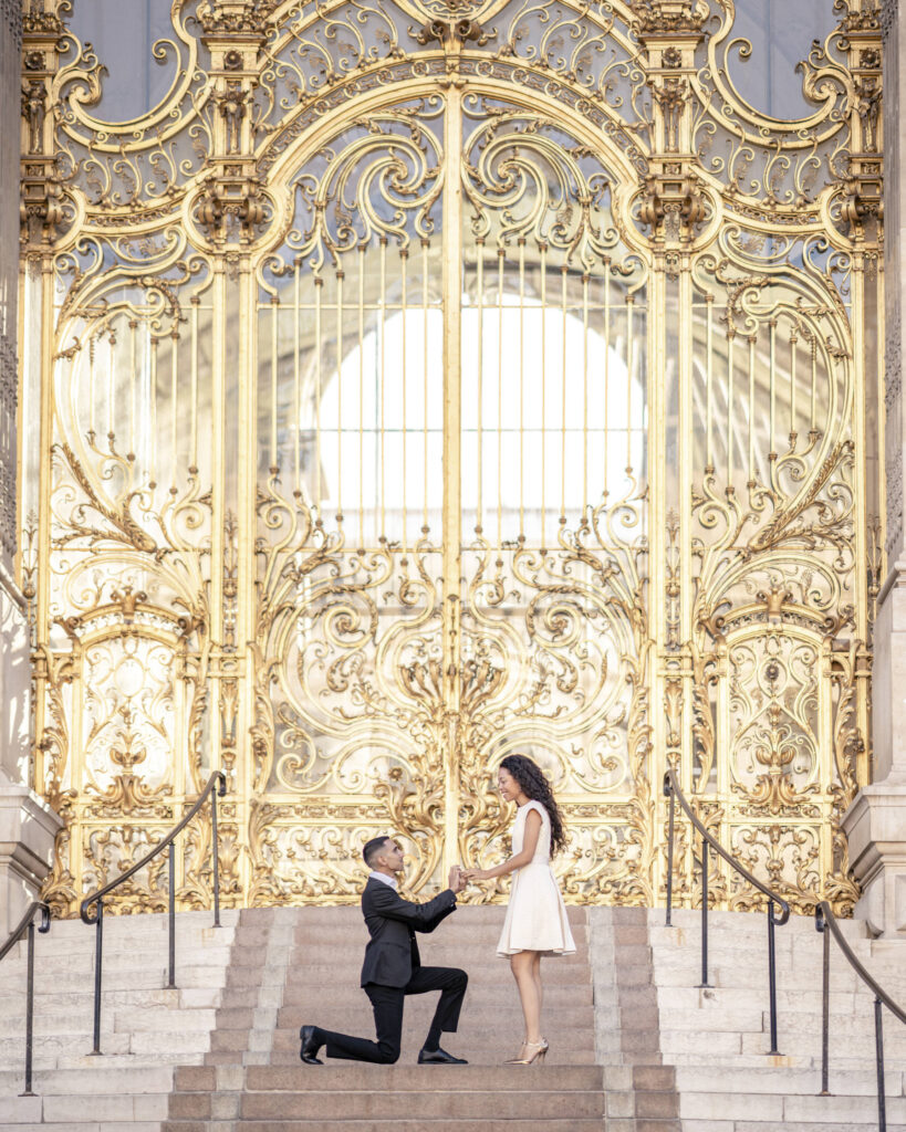 Man down on one knee and proposing to his girlfriend on the steps of the Petit Palais in front of the golden gates.