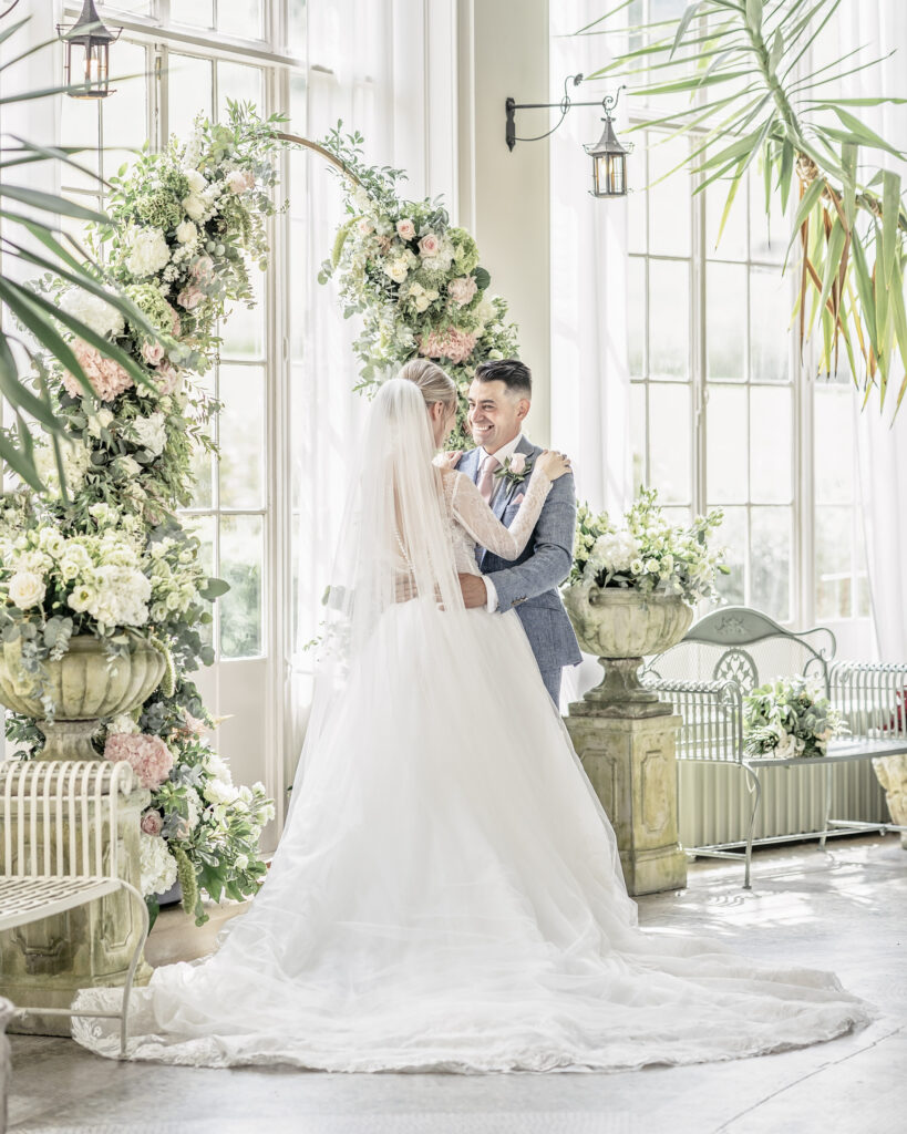 Bride and groom posing together by a large floral arch in the Winter Garden room at Hawkstone Hall in Shropshire.