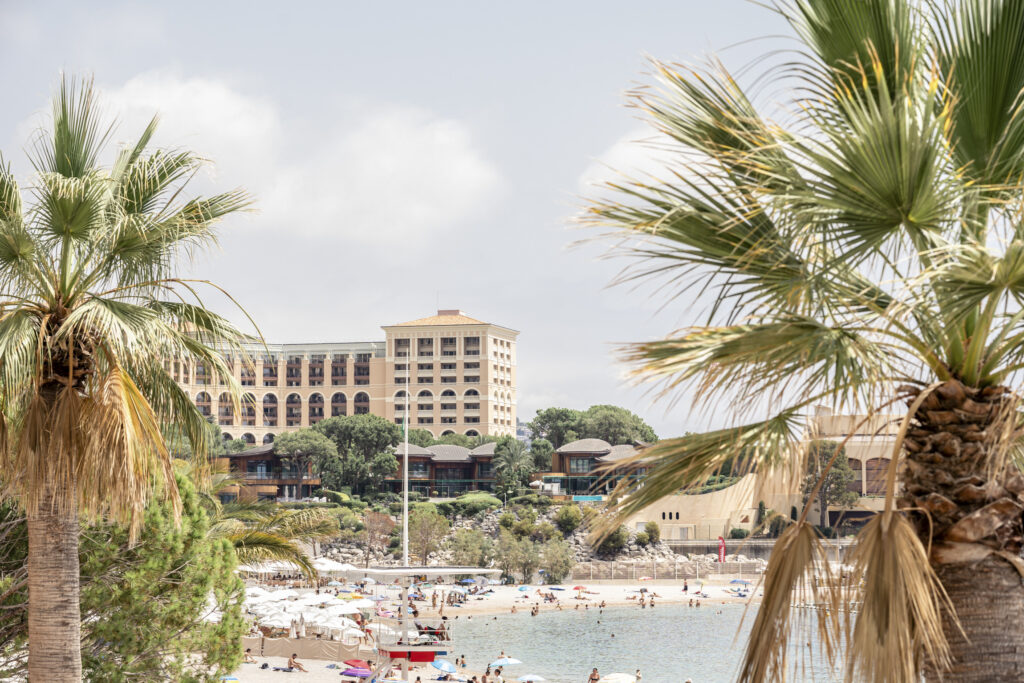 A landscape photo of Monaco with a 5-star hotel, beach packed with sunbathers and palm trees.