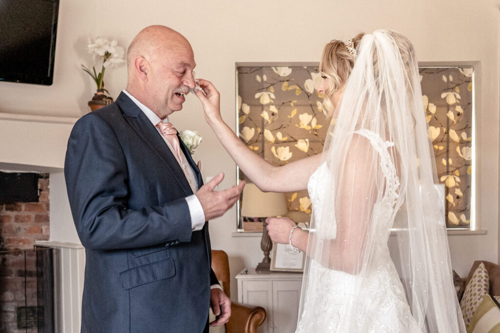 Bride wiping away her father's tears with a tissue in touching emotional moment after he sees his bride daughter for the first time in her wedding dress.