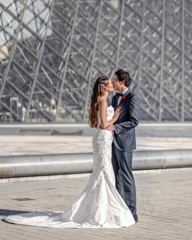 Bride and groom hugging and kissing in front of the glass pyramid at the Louvre museum in Paris