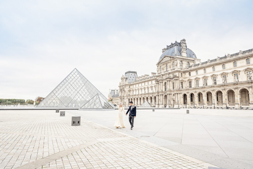 A bride and groom walking hand in hand in front of the large Louvre pyramid in Paris. The bride is raising her bouquet in the air in celebration.