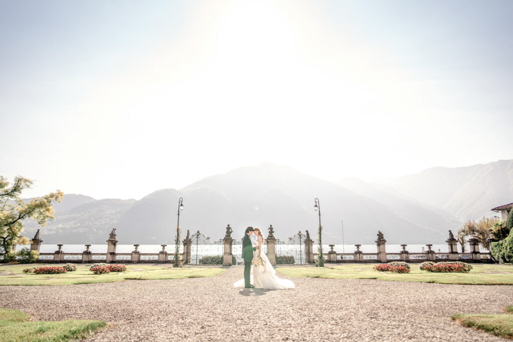 A bride and groom posing together in the front gardens of Villa Sola Cabiati with Lake Como and the mountains in the background.