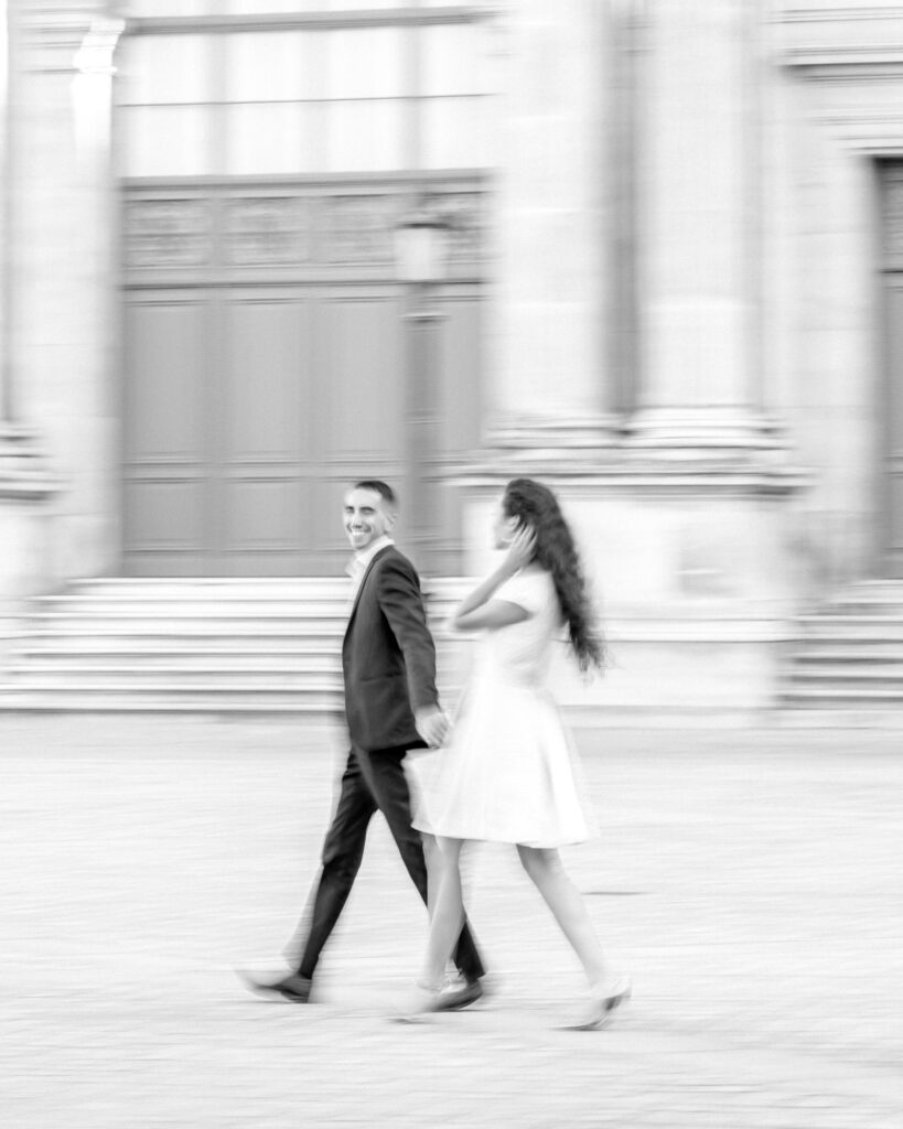 A black and white, movement blur photo of a bride and groom to be walking hand in hand next to the Louvre Pyramid in Paris for their engagement shoot.