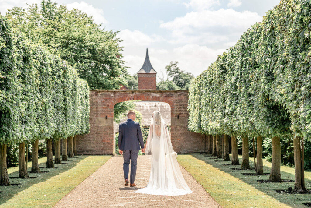 Bride and groom walking hand in hand down the tree lined path at Combermere Abbey.