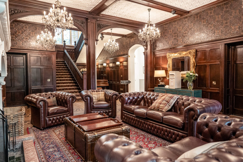 The front reception room at Tyn Dwr Hall with leather chesterfield sofas, chandeliers, fireplace, and staircase.