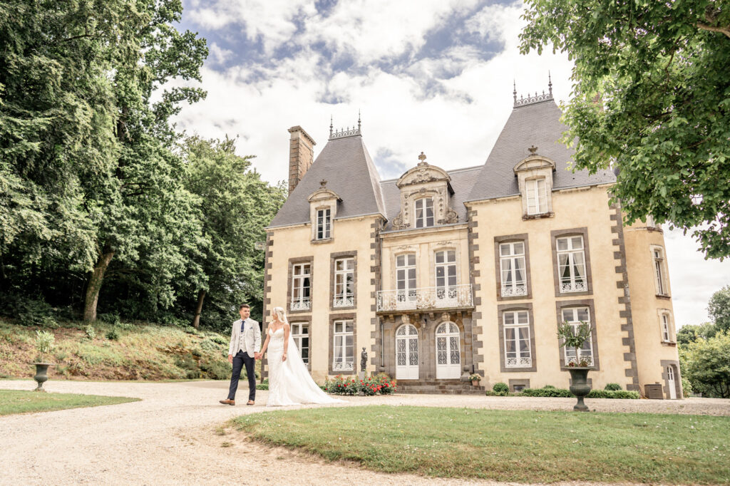 A bride and groom walking hand in hand and looking at each other in front of Chateau du Grand Val wedding venue in Brittany, 