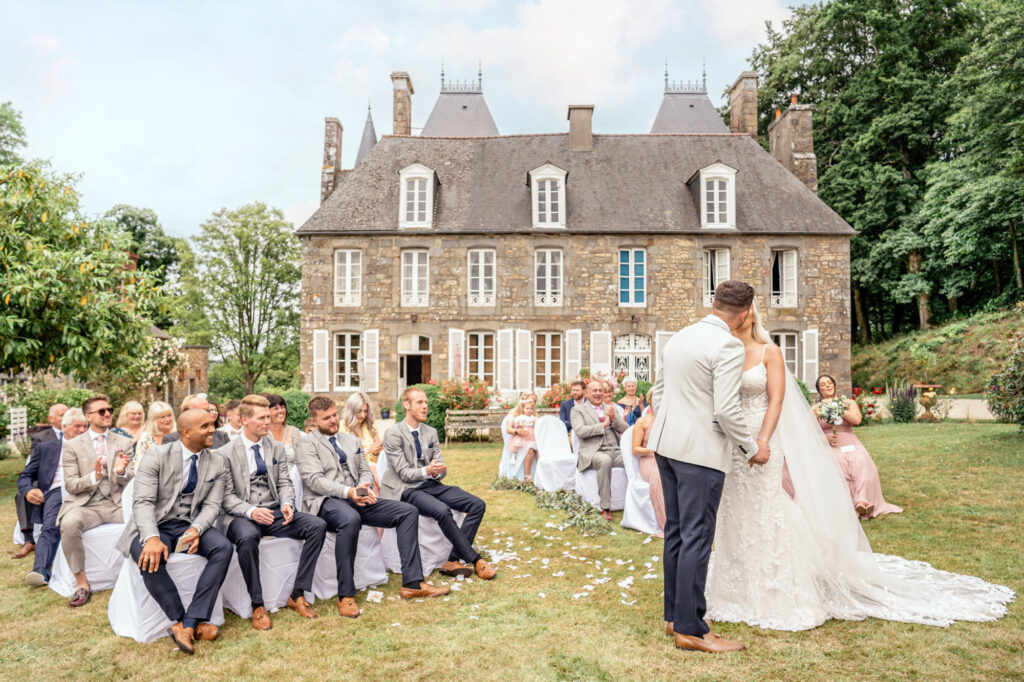 The first kiss while guests clap the bride and groom on during an outside wedding in the garden at Chateau du Grand Val in Brittany, France. The Chateau can be seen in the background.