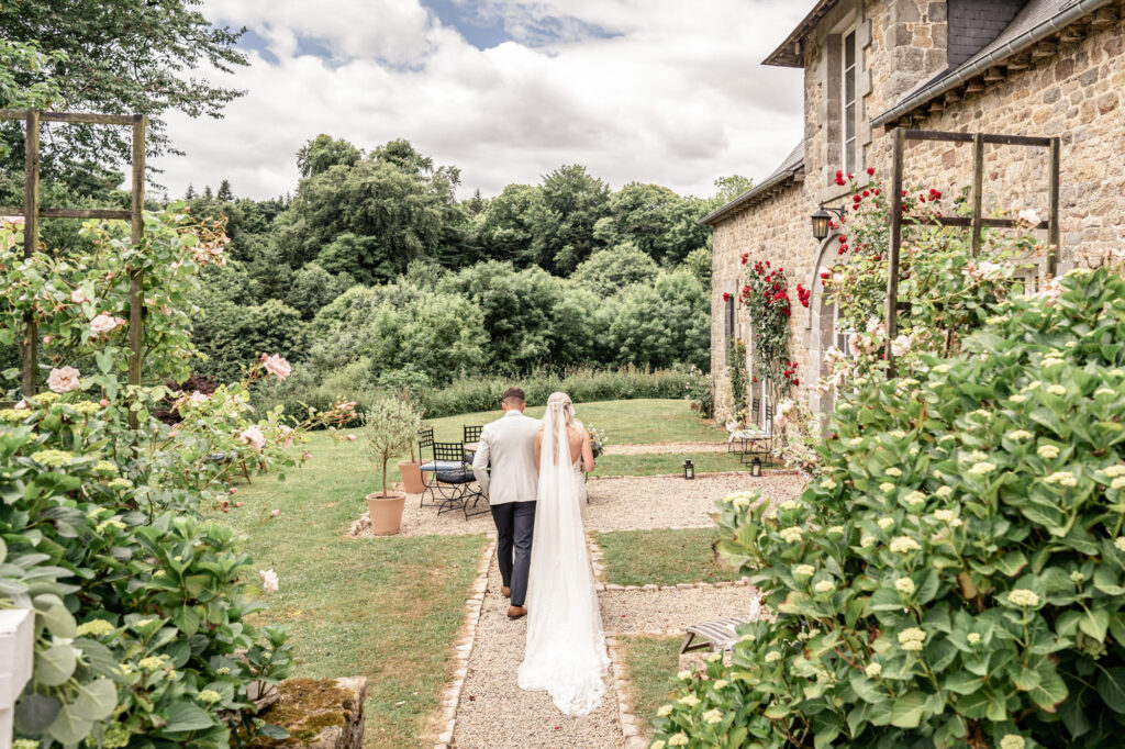 A bride and groom photographed from behind walking through the garden to the Rose House (old Cider House) at Chateau du Grand Val wedding venue in Brittany, France. 