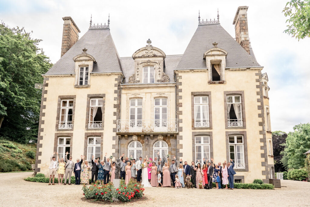 A full group photo of a large wedding party waving to the camera in front of Chateau du Grand Val in Brittany, France. 